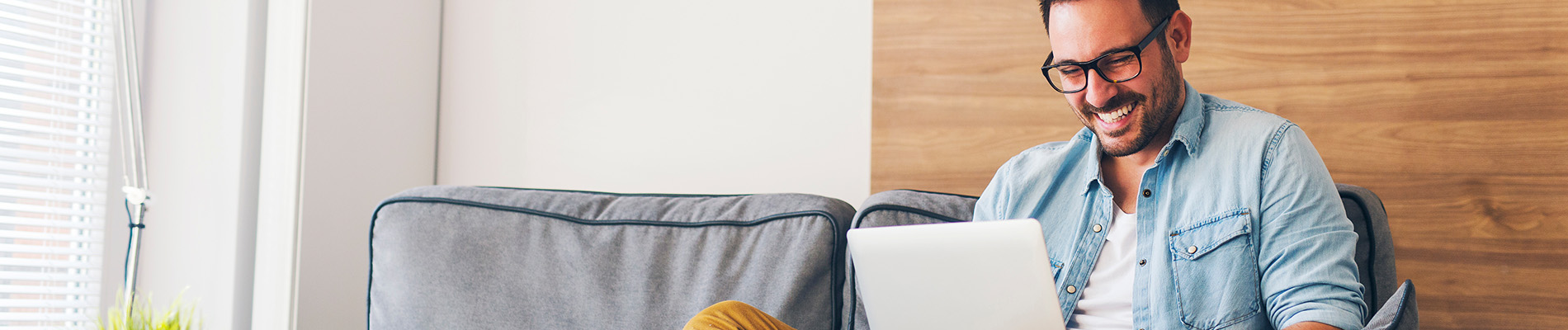 The image shows a man sitting at a desk with a laptop, wearing glasses and smiling, with a blurred background suggesting an office environment.