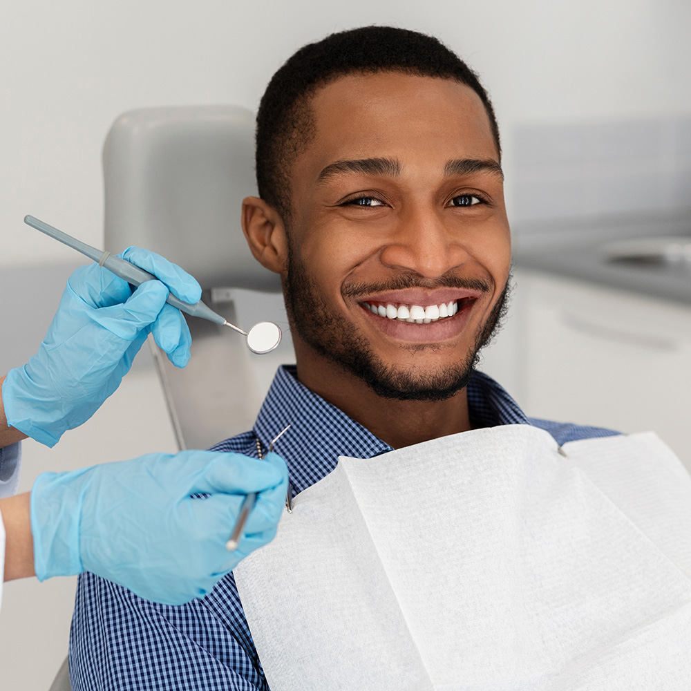 This image shows a person sitting in a dental chair, smiling at the camera while wearing a blue surgical mask, with their mouth open as if they are receiving dental care, and there s a dental hygiene tool visible near them.